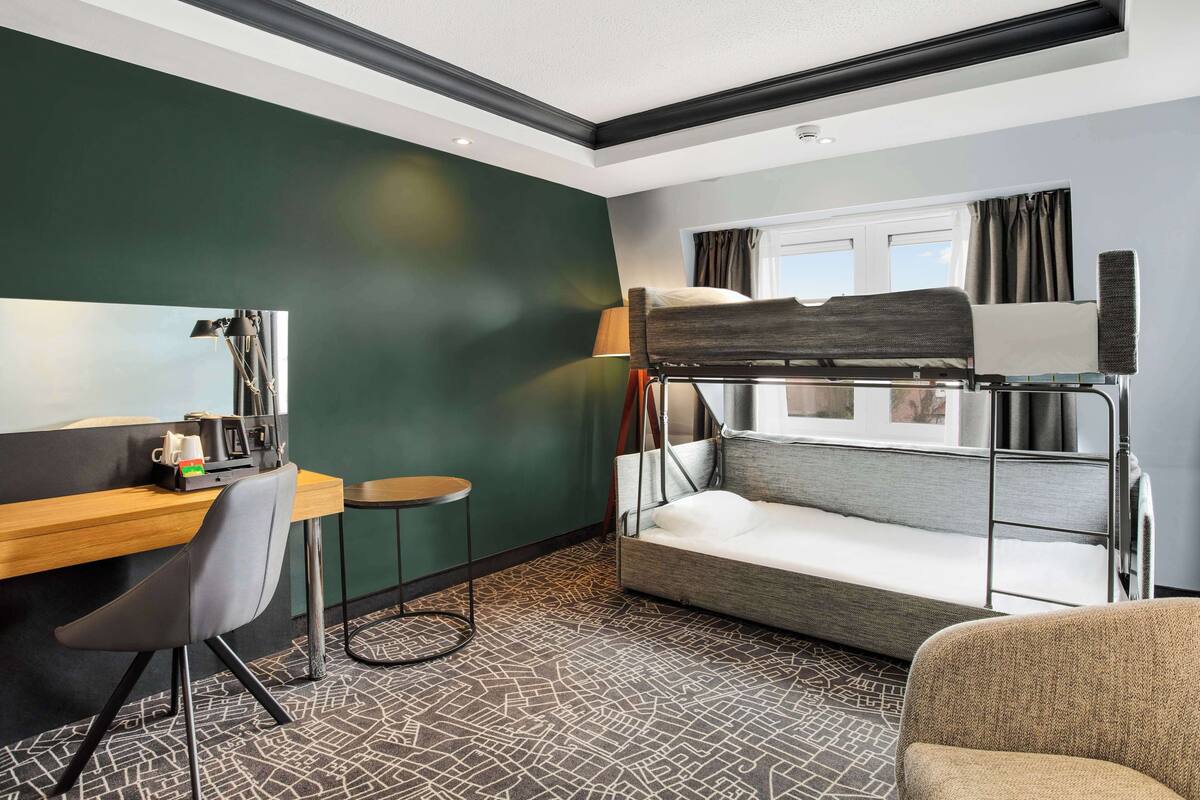 A compact hotel room with a bunk bed, a desk with a chair, a small table, and a lamp.