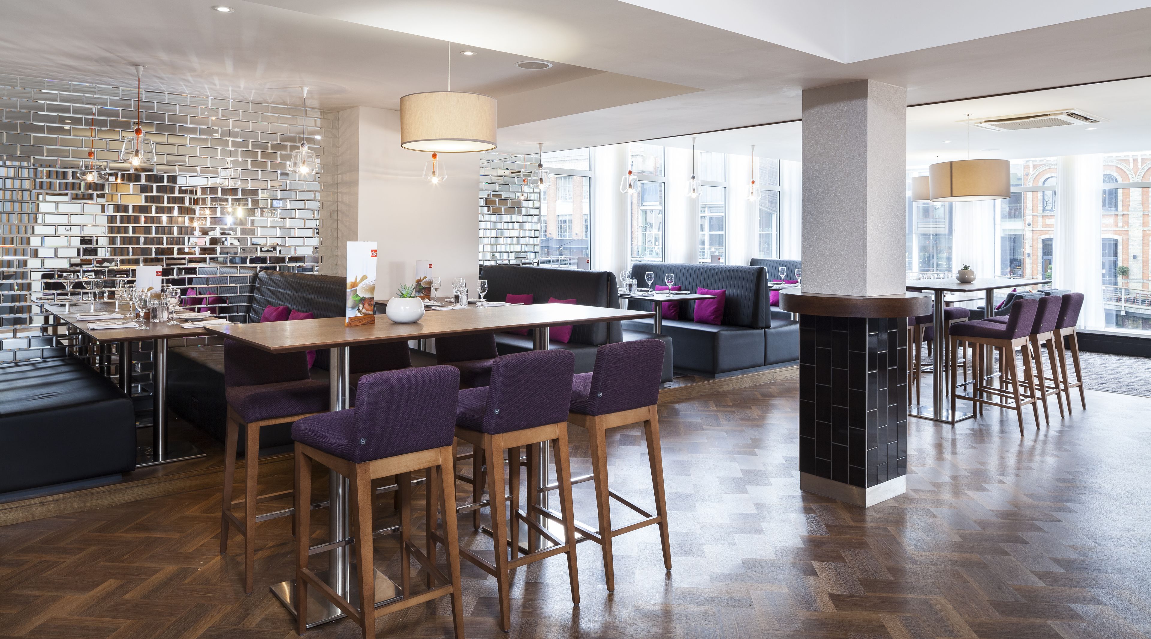 A modern dining area with a brick wall, wooden tables, purple chairs, and large windows offering city views.