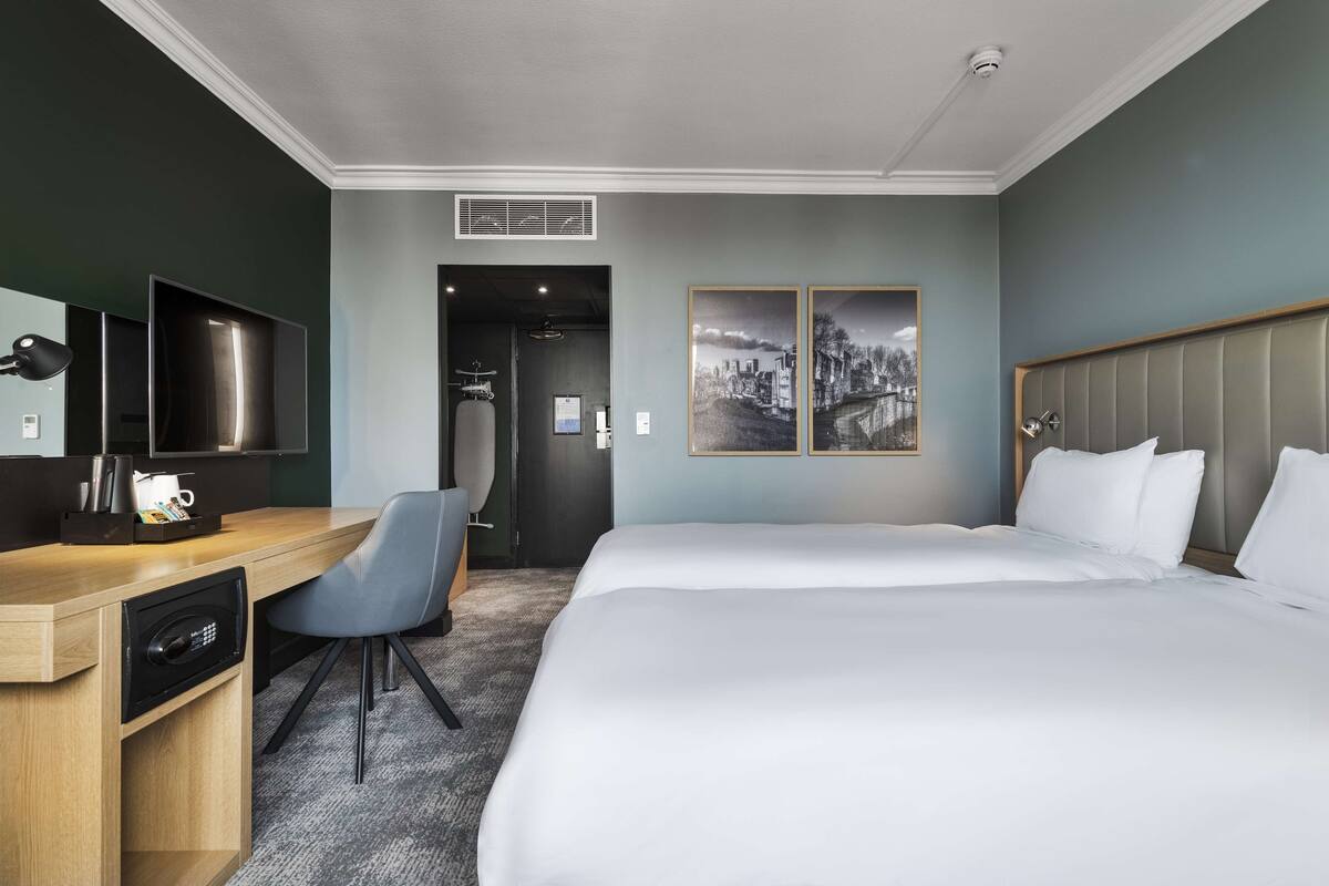 A hotel room with a bed, desk, chair, TV, and artwork on the wall.