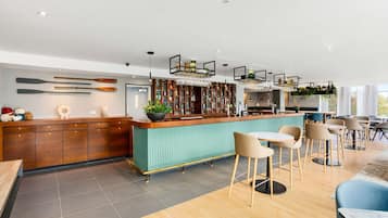 Una barra moderna con mostrador verde azulado, armarios de madera y una variedad de botellas. Se ven varias sillas y una mesa en primer plano.