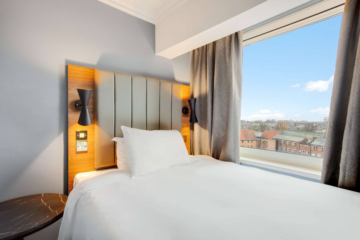 A hotel room with a large bed, a wooden headboard, a nightstand, a lamp, and a view of buildings through a window.