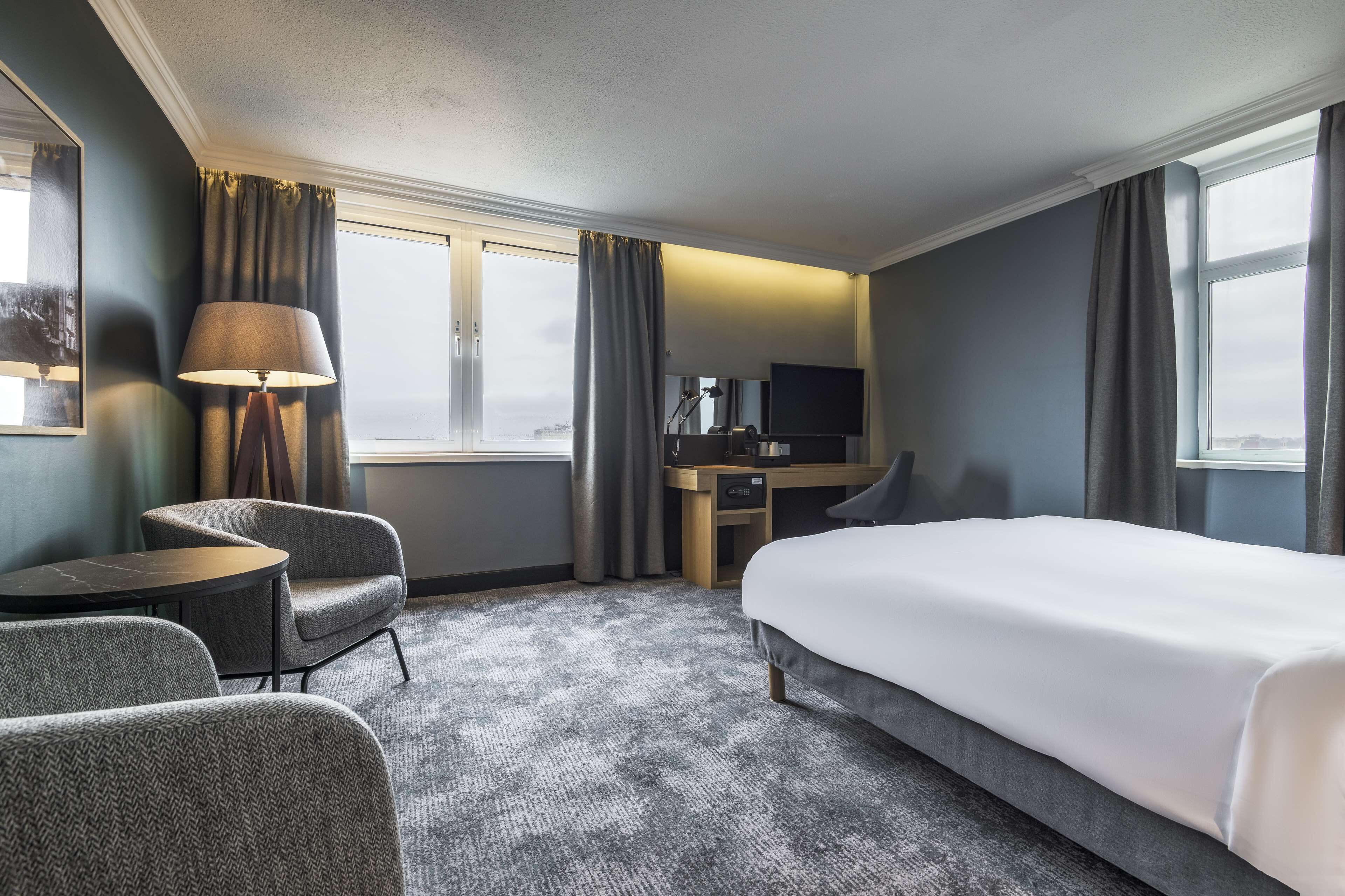 A hotel room with a bed, a desk, a chair, a lamp, and a TV.