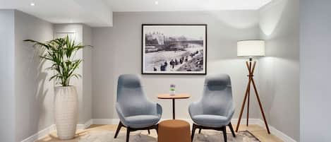 A modern room with two grey armchairs, a small round table, a large potted plant, a framed black and white photo on the wall, and a floor lamp.