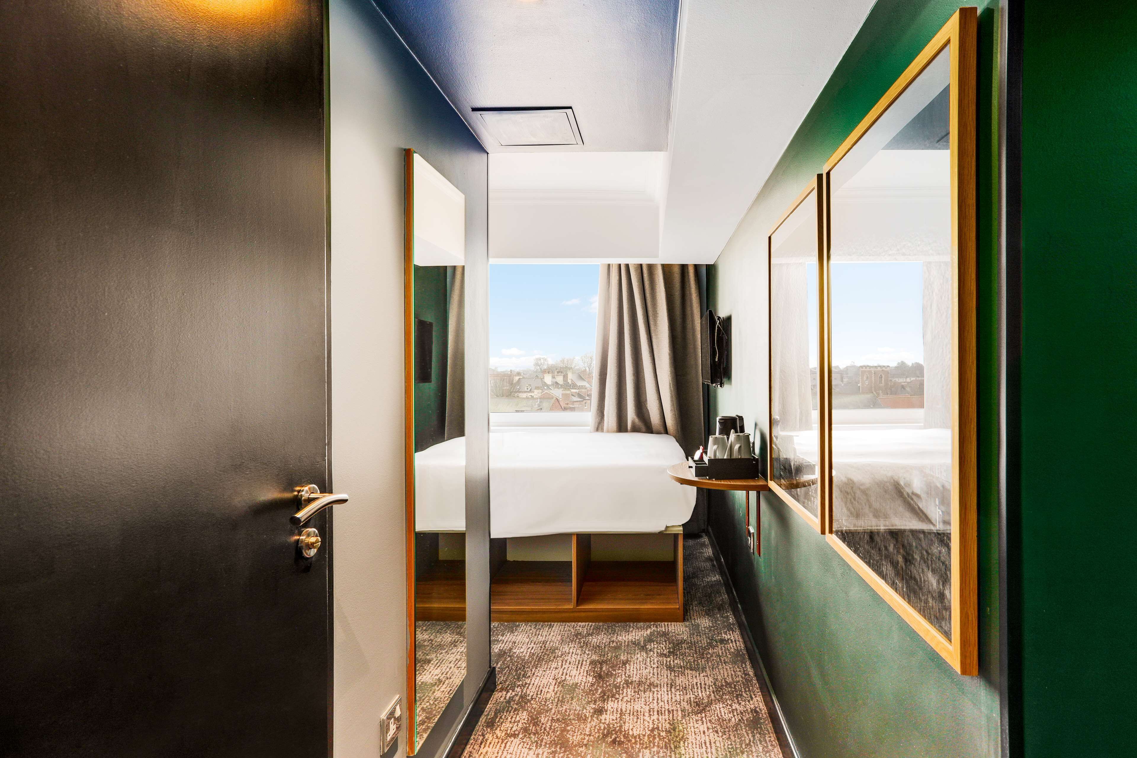 A hotel room with a bed, a small table with a lamp, a window with curtains, and a view of the city.