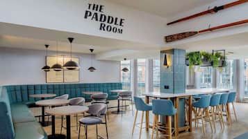 A modern interior with a green padded bench seating area, round tables, and blue chairs. A bar area with stools is visible, and there are wooden paddles hanging on the wall.