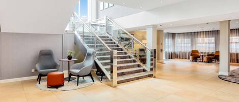 A spacious lobby with a central staircase, seating area with chairs and a small table, and large windows allowing natural light.