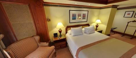 Una habitación de hotel con una cama grande, un sillón a rayas, dos mesitas de noche con lámparas y un cuadro enmarcado en la pared.
