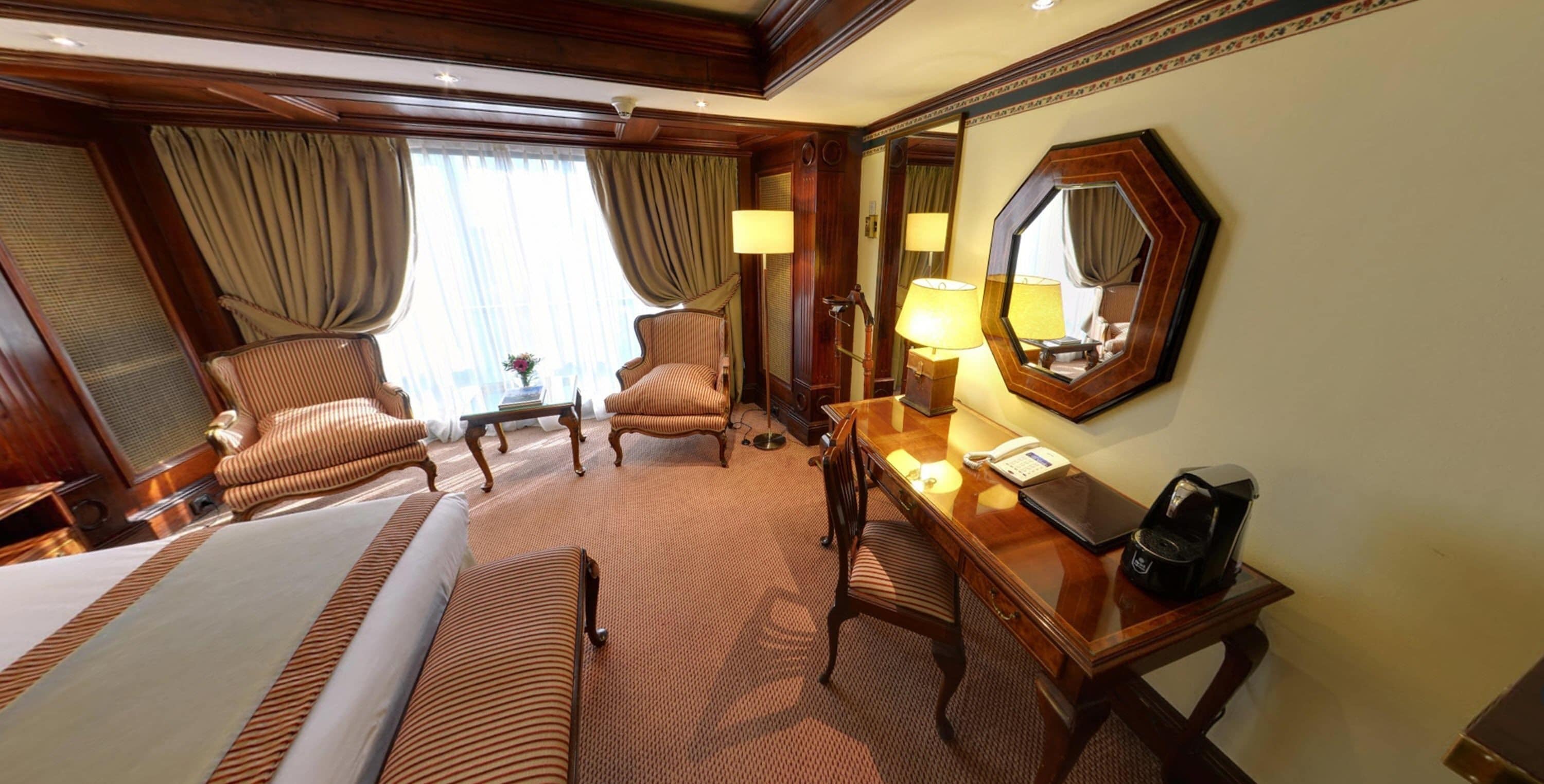 Habitación de hotel con una cama grande, dos sillones, un escritorio con lámpara, un espejo y piso alfombrado.