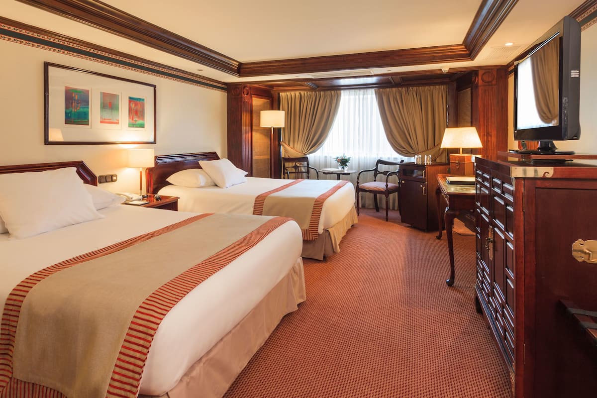 A hotel room with two beds, a desk with a lamp, a chair, a television, and a window with curtains.
