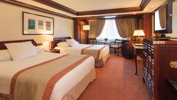 A hotel room with two beds, a desk with a lamp, a chair, a television, and a window with curtains.