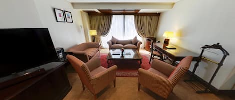 Habitación de hotel con televisión de pantalla plana, un sofá, dos sillones a rayas, una mesa de centro, una caminadora y un ventanal con cortinas.