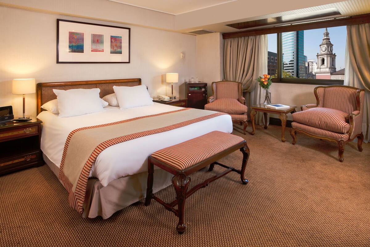 A hotel room with a large bed, a bench, two armchairs, a small table with a vase, and a city view through the window.