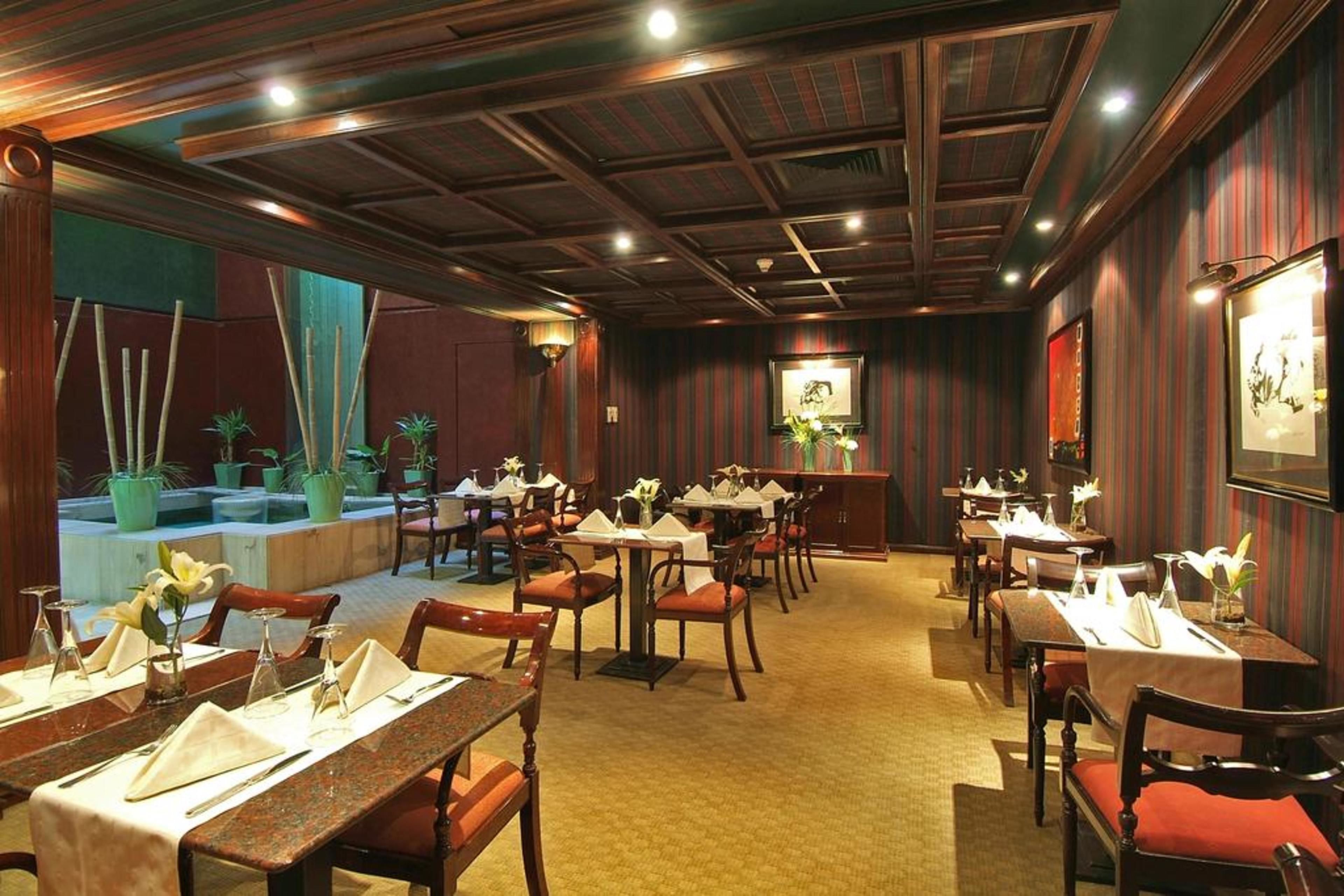 Restaurante com vigas de madeira no teto, mesas com toalhas brancas e cadeiras vermelhas, e plantas em vasos.