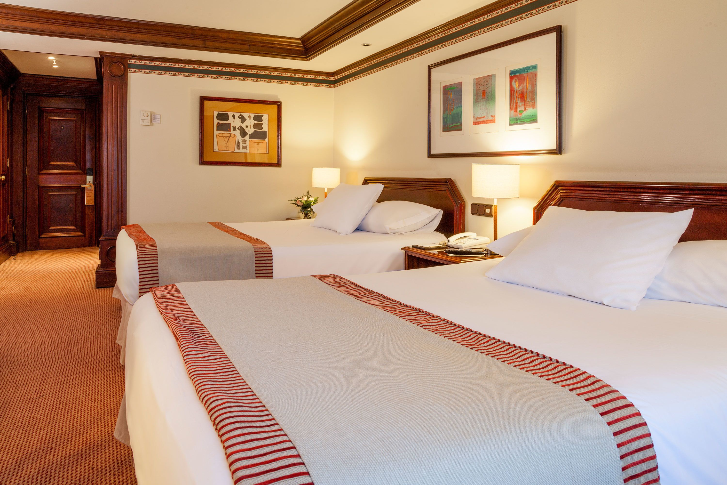 A hotel room with two beds, each with a red-striped bedspread, a wooden headboard, and a nightstand with a lamp.