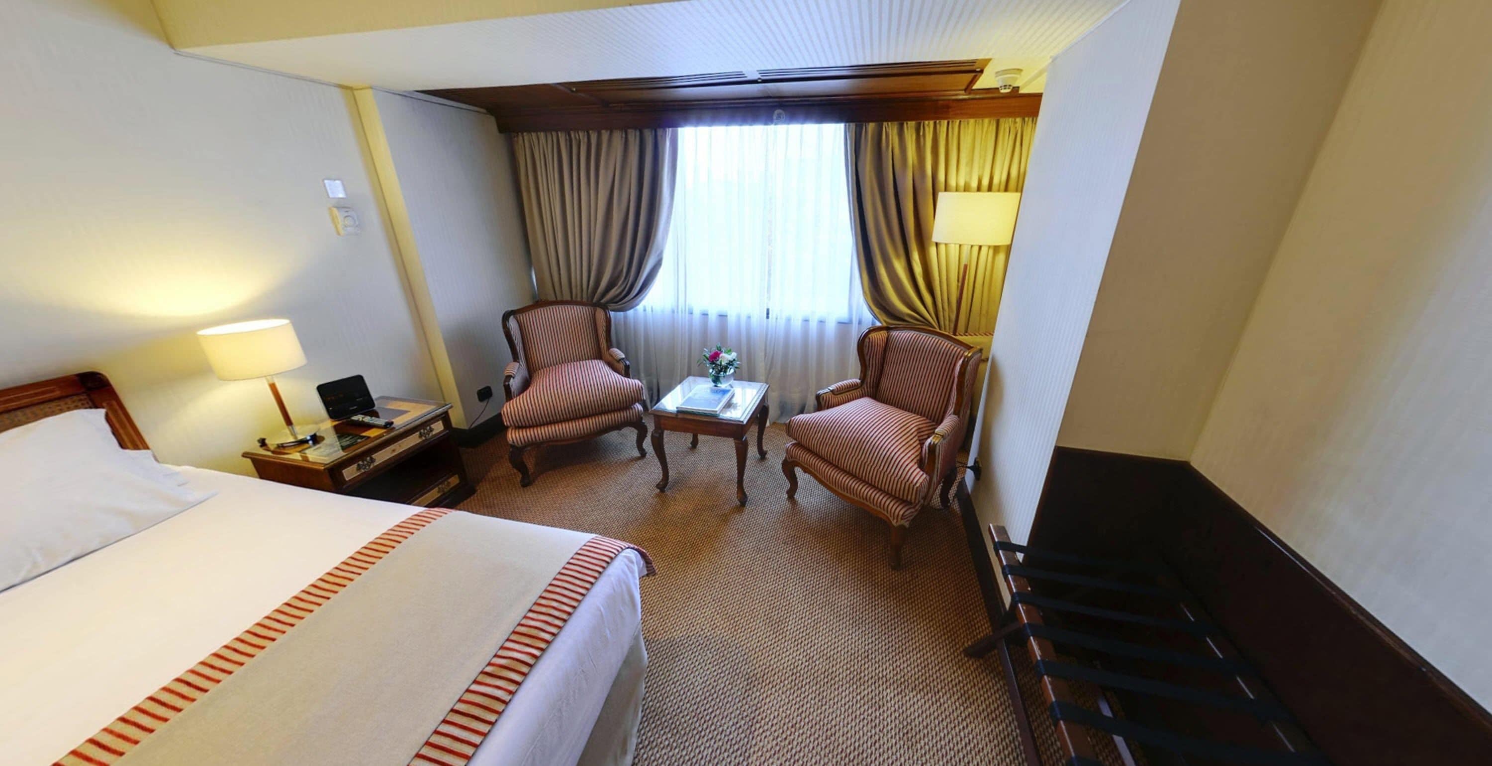 A hotel room with a large bed, two armchairs, a small table, and a desk with a lamp.