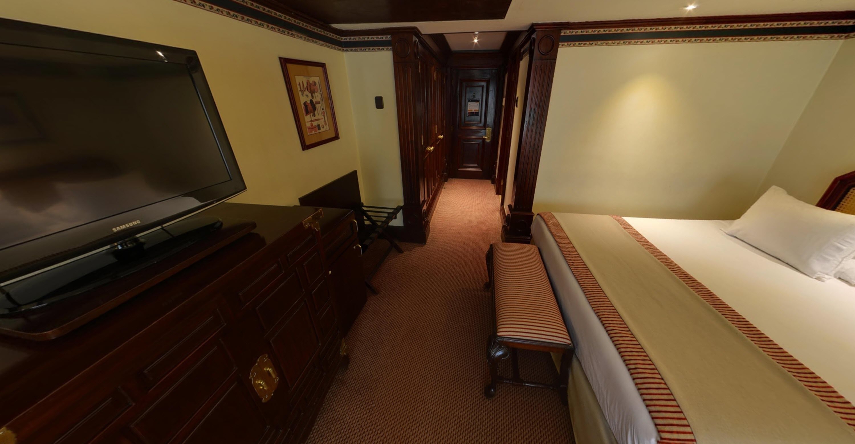 Une chambre d’hôtel dotée d’un grand téléviseur à écran plat, d’une commode en bois, d’un banc, d’un lit avec une literie à rayures et d’un couloir menant à une autre chambre.