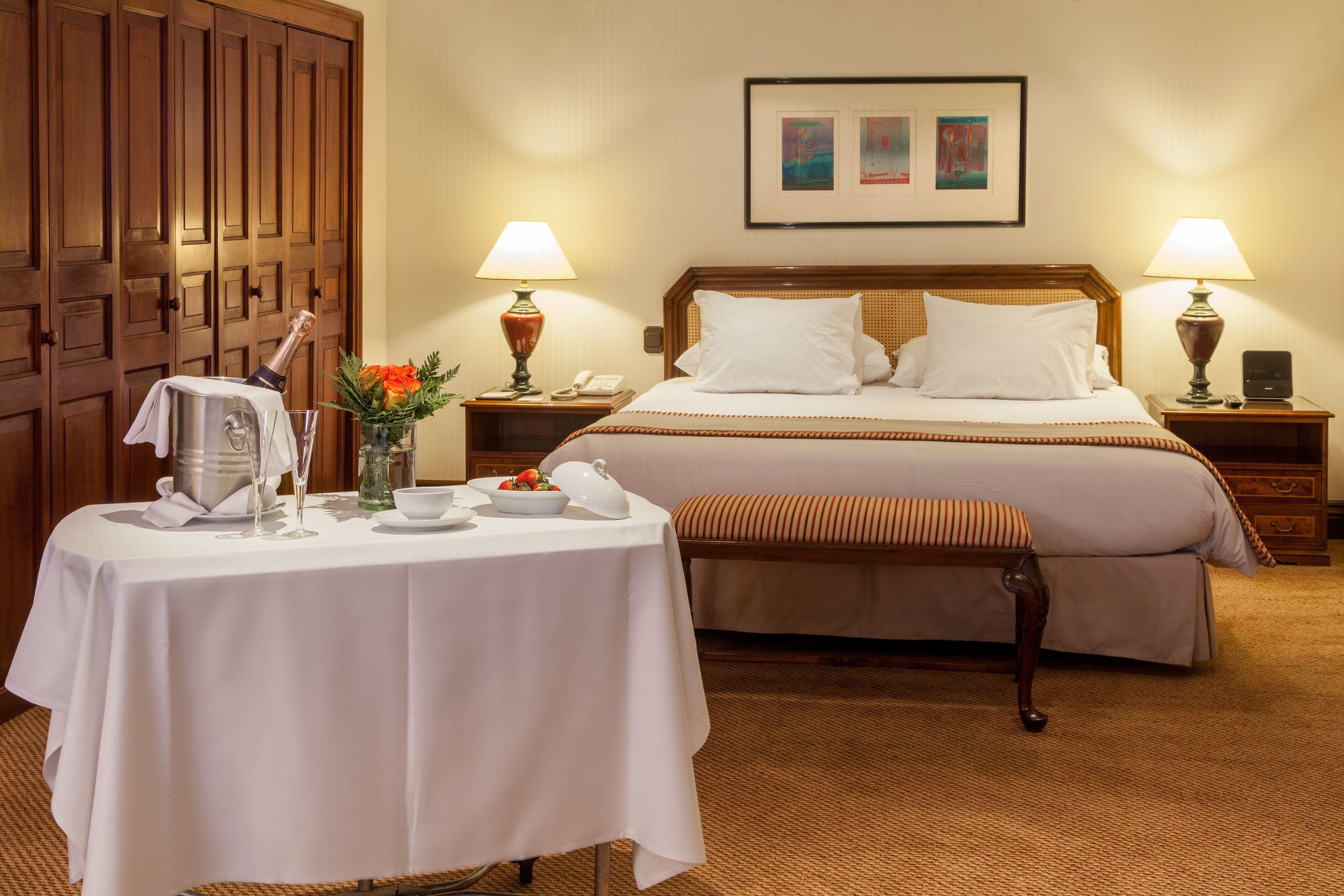 A hotel room with a bed, bedside tables, a small table with a bottle of champagne, a vase with flowers, and a framed picture on the wall.