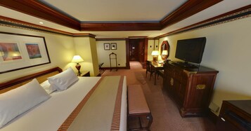 A hotel room with a large bed, a desk with a chair, a television, and framed artwork on the wall.