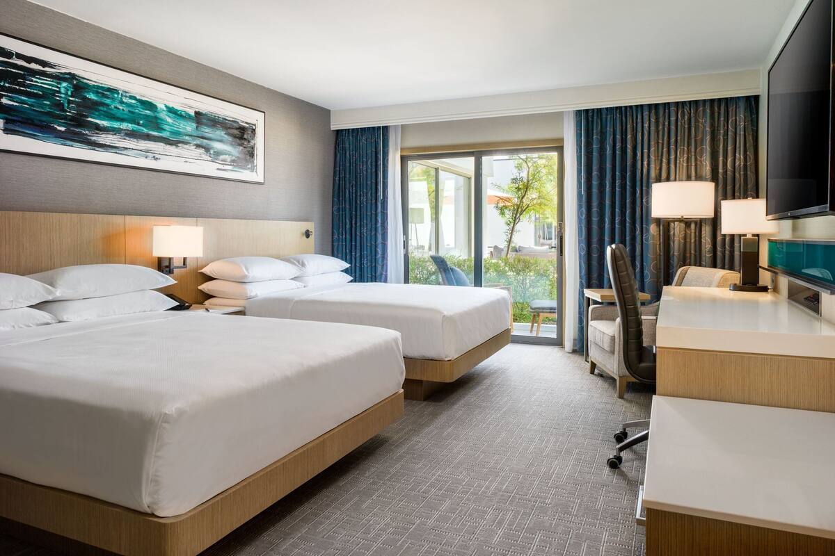A hotel room with two beds, a desk, a chair, a TV, and a large window with curtains.