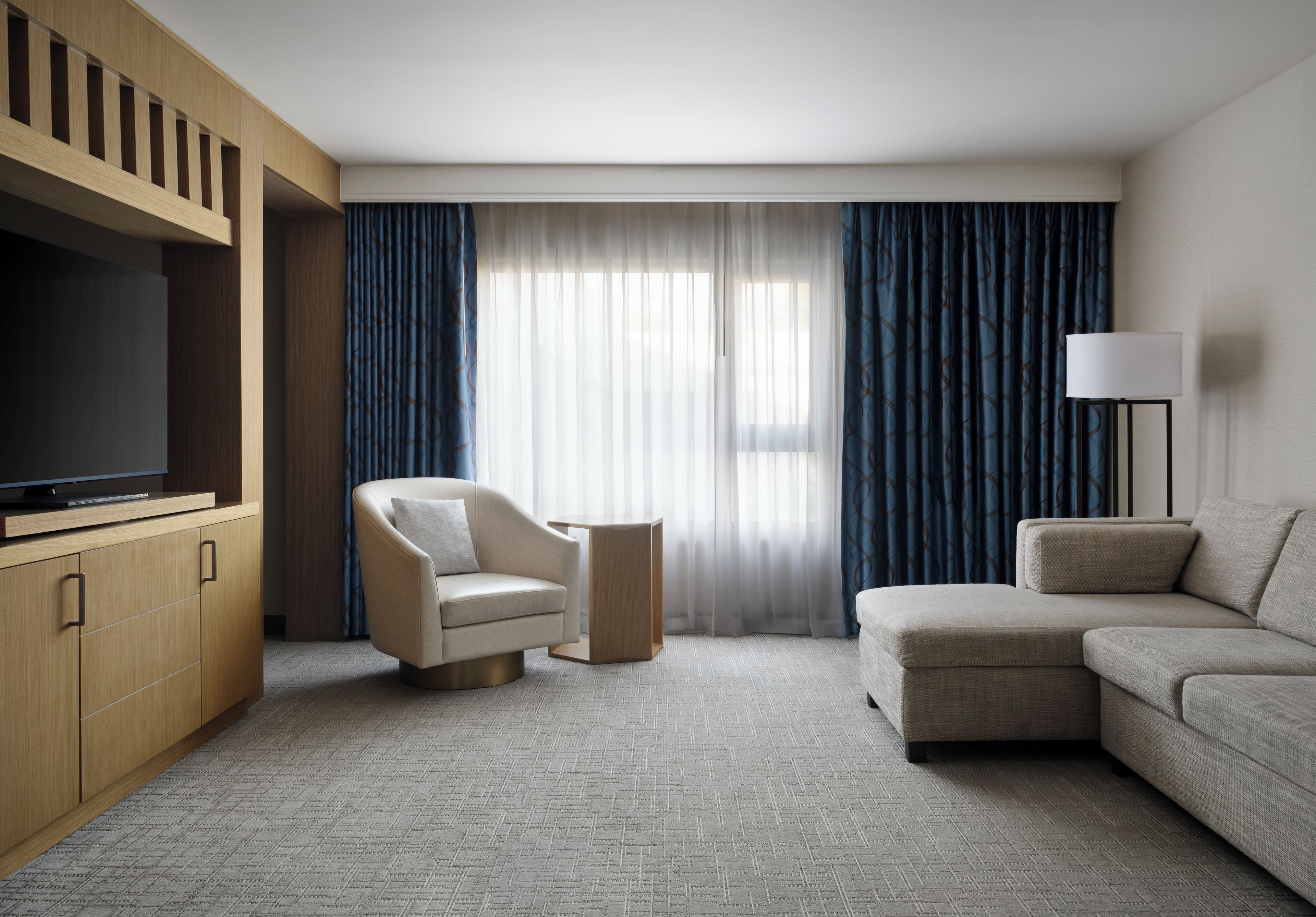 A hotel room with a flat-screen TV, a sofa, a chair, and a large window with curtains.