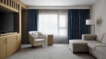 A hotel room with a flat-screen TV, a sofa, a chair, and a large window with curtains.