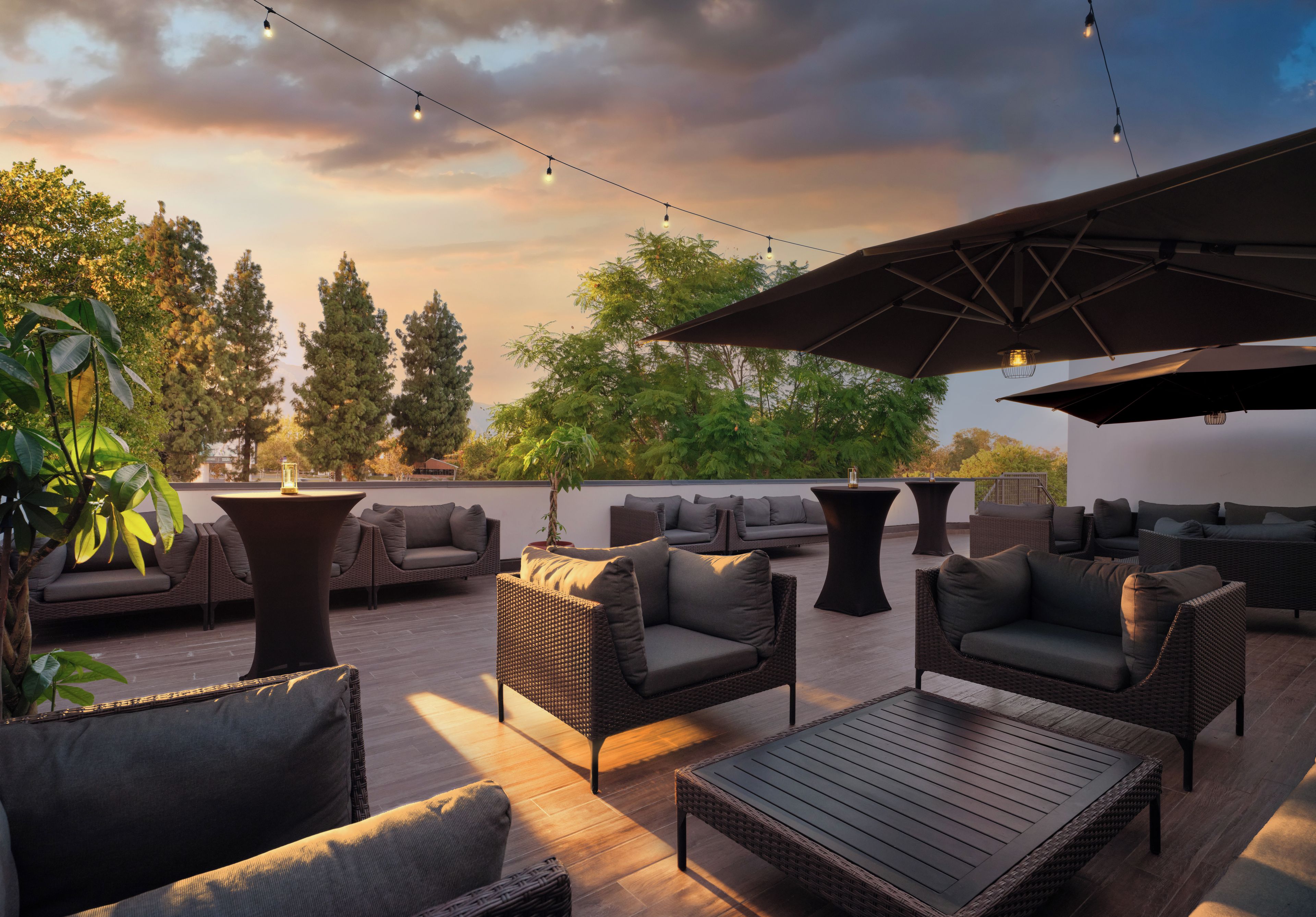 Un área de estar al aire libre con mobiliario de mimbre, una mesa y luces colgantes, con árboles y un cielo de atardecer de fondo.
