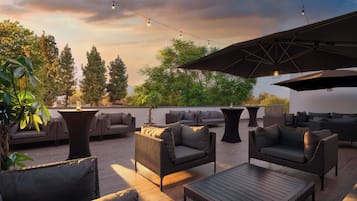 Un área de estar al aire libre con mobiliario de mimbre, una mesa y luces colgantes, con árboles y un cielo de atardecer de fondo.