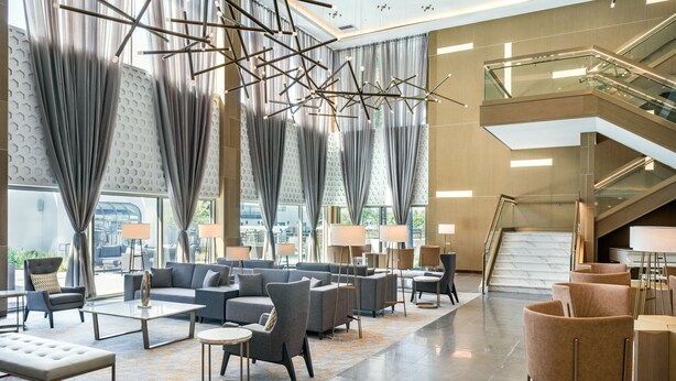 A spacious lobby with a modern design, featuring a seating area with sofas and armchairs, a staircase, and large windows with curtains. - Delta Hotels by Marriott Ontario Airport (Ontario)