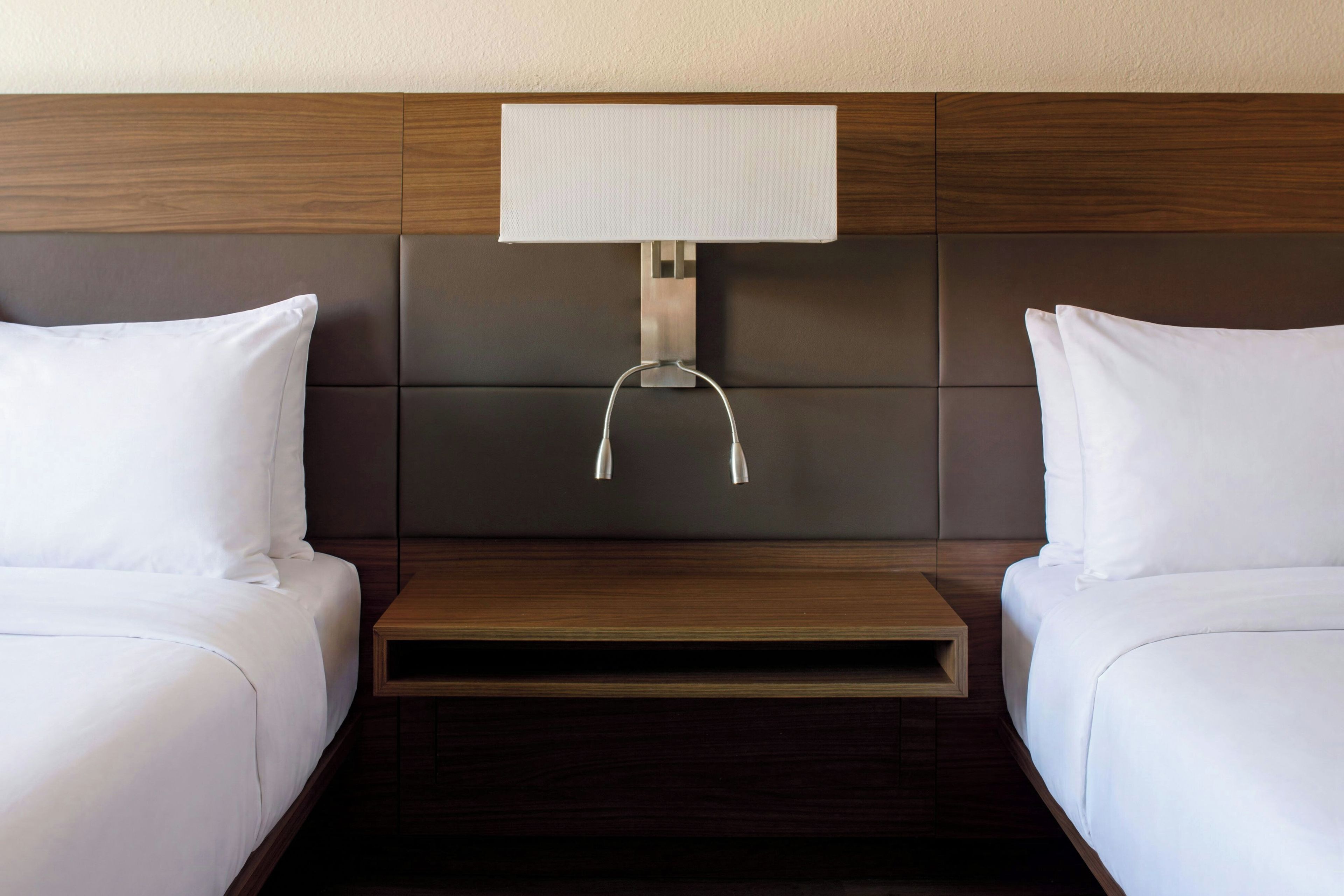 A hotel room with two beds, a nightstand, and a wall-mounted lamp.