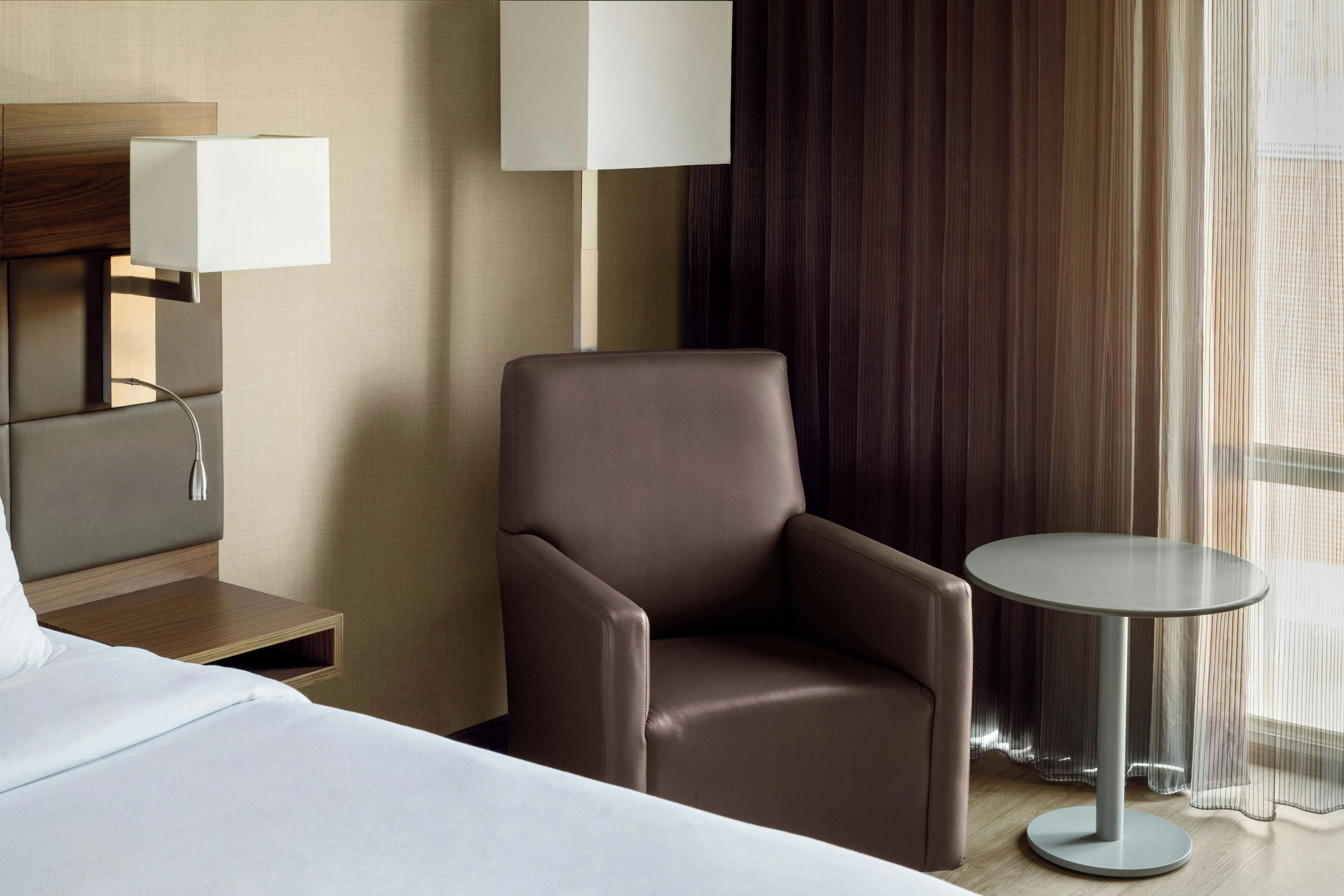 A hotel room with a bed, a chair, a small table, a lamp, and a window with curtains.