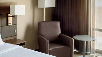 A hotel room with a bed, a chair, a small table, a lamp, and a window with curtains.