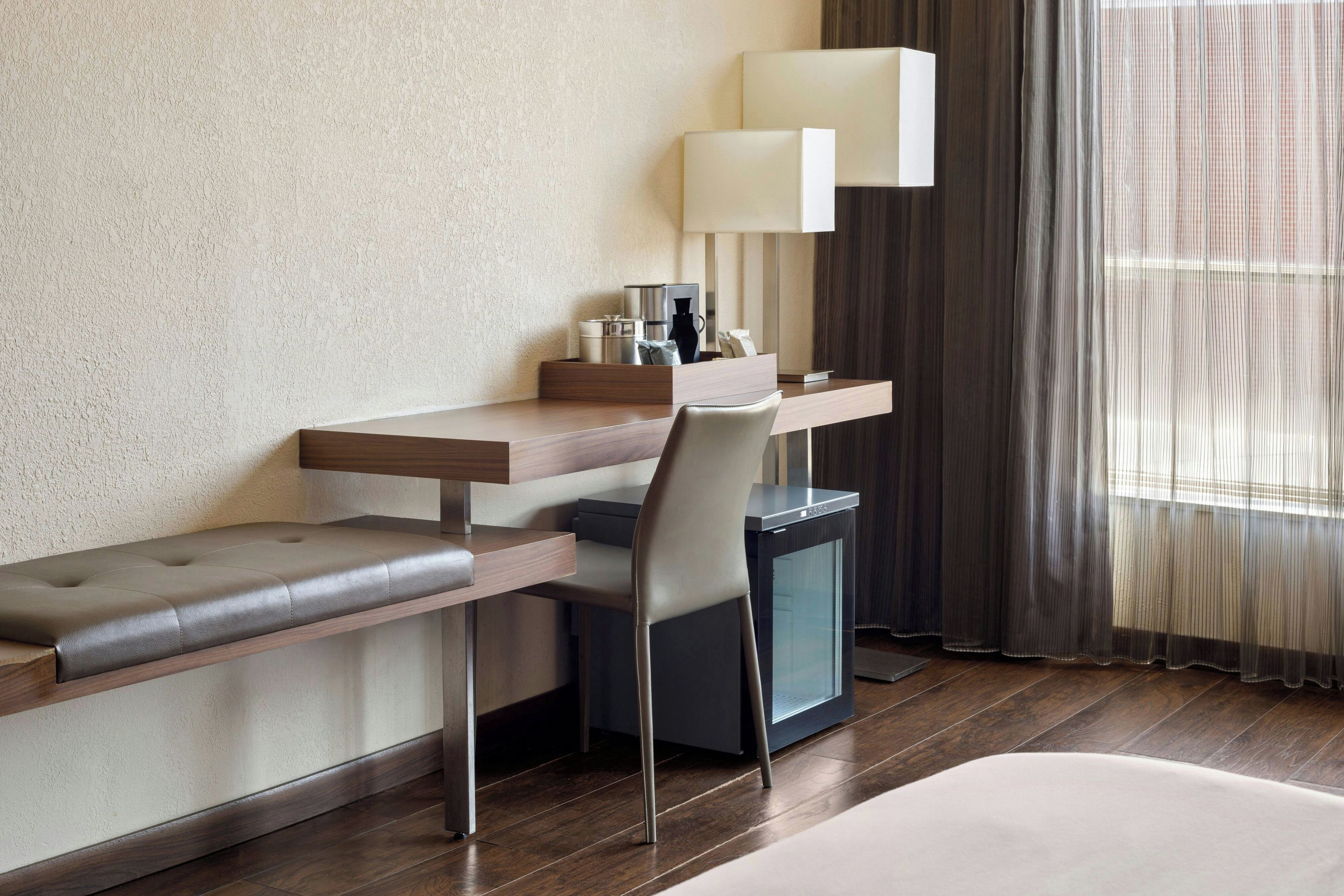 A hotel room with a wooden desk, a cushioned bench, a chair, a small cabinet, and a bed.