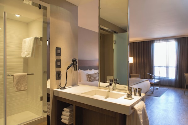 Un baño de hotel con cabina de ducha de vidrio, un lavabo con espejo y una cama visible a través del espejo.