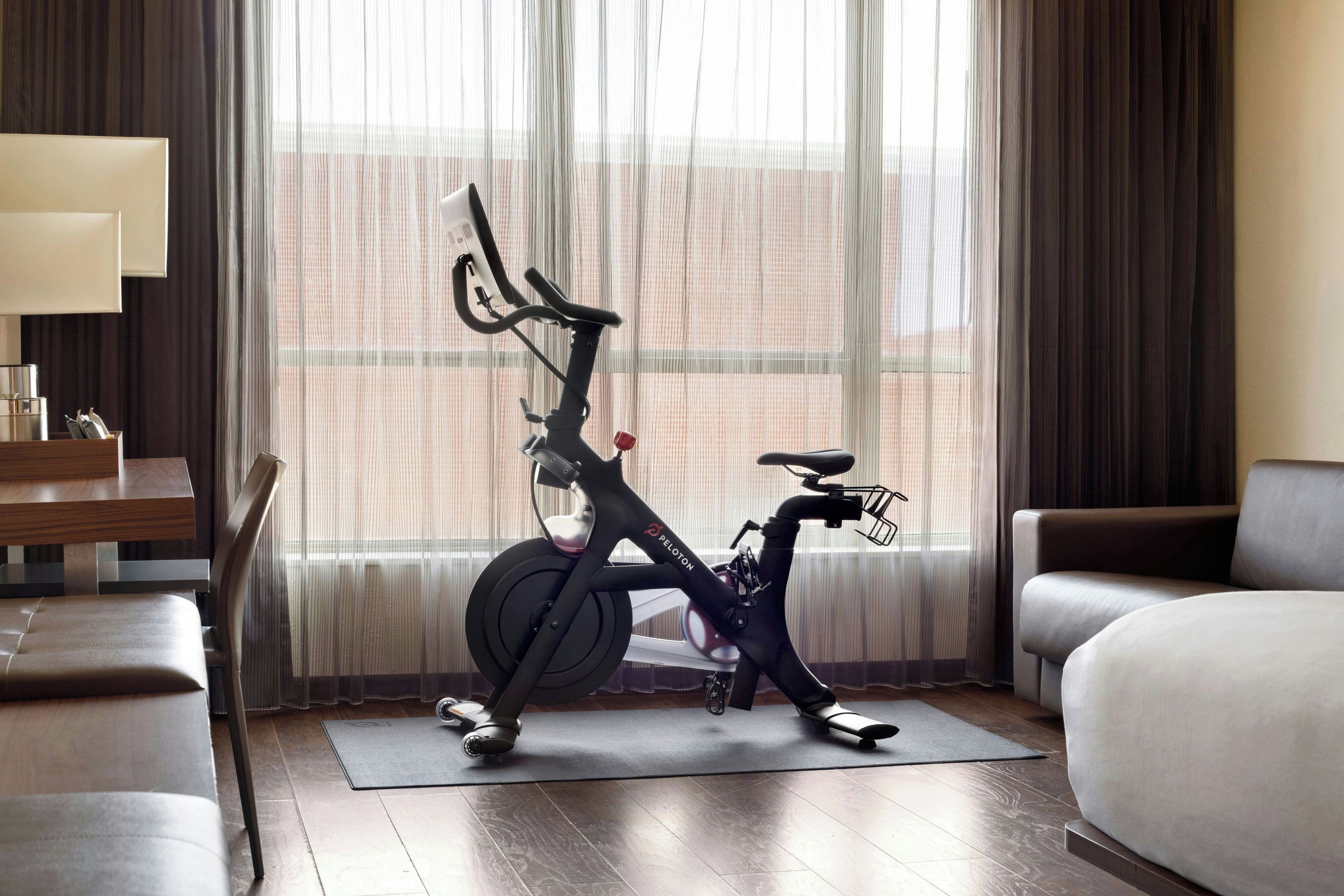 A hotel room with a stationary bike, a bed, a desk, and a window with curtains.