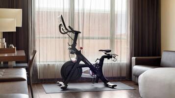 A hotel room with a stationary bike, a bed, a desk, and a window with curtains.