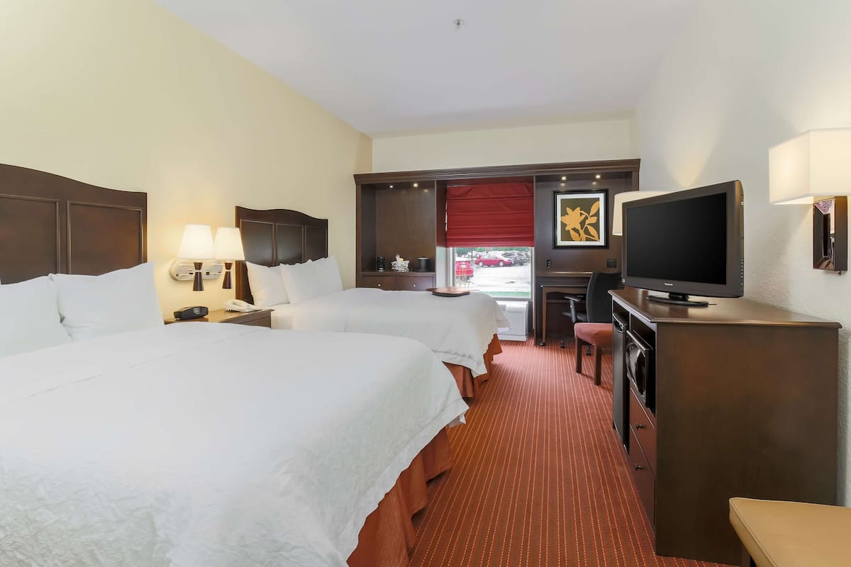 A hotel room with two beds, a desk, a television, and a window with a red blind.