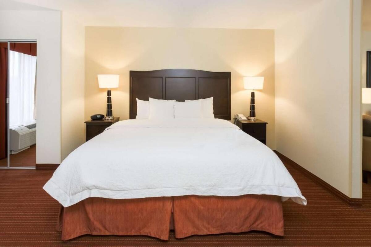 A hotel room with a large bed, two bedside tables with lamps, a mirror, and a view of a window with curtains.