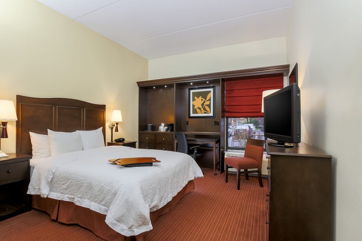A hotel room with a large bed, a desk, a chair, a television, and a window with a red curtain.