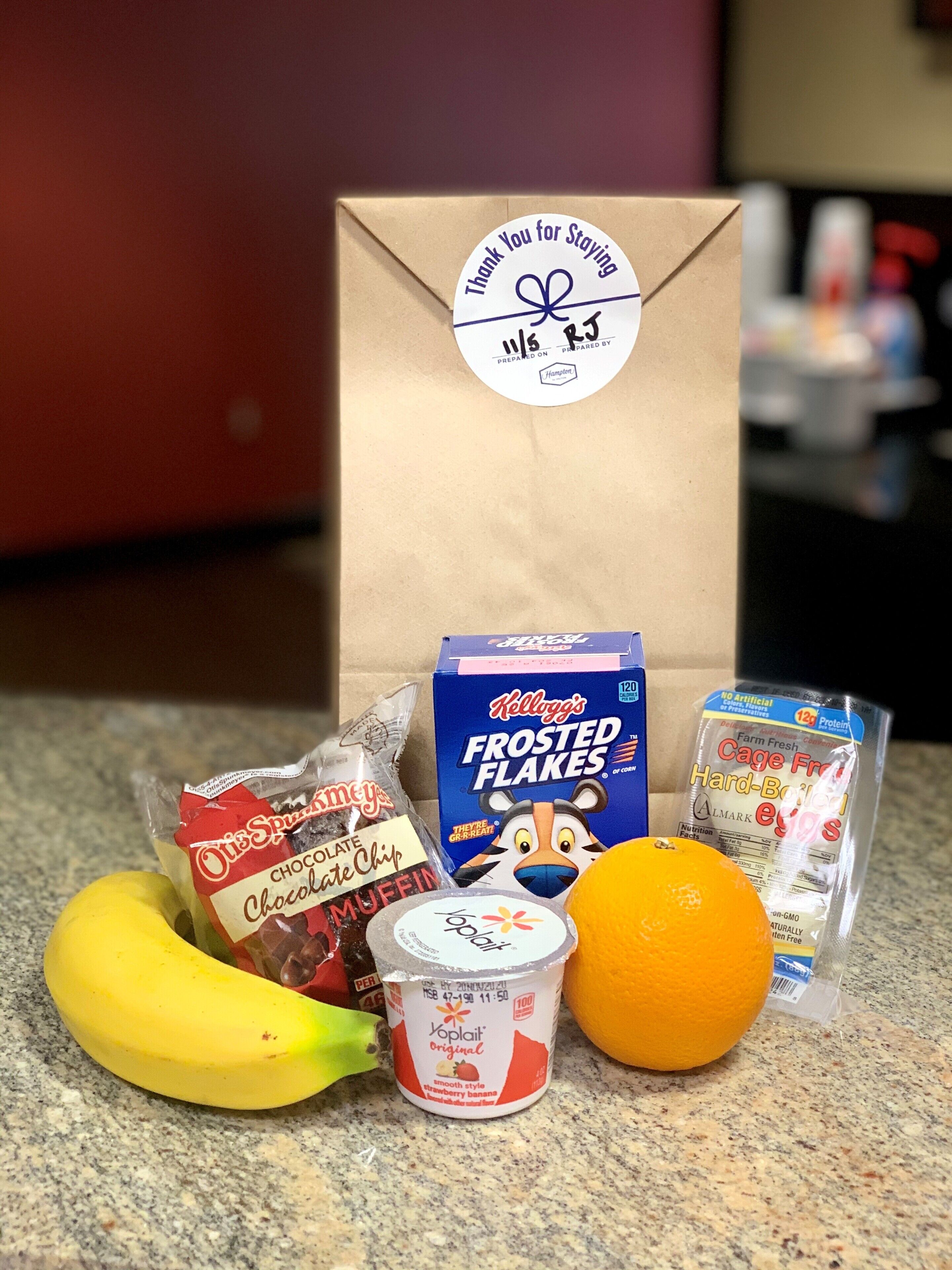 A gift bag with a thank you note, a box of Kellogg's Frosted Flakes, a banana, a cup of yogurt, a bag of chocolate chips, and a package of eggs.