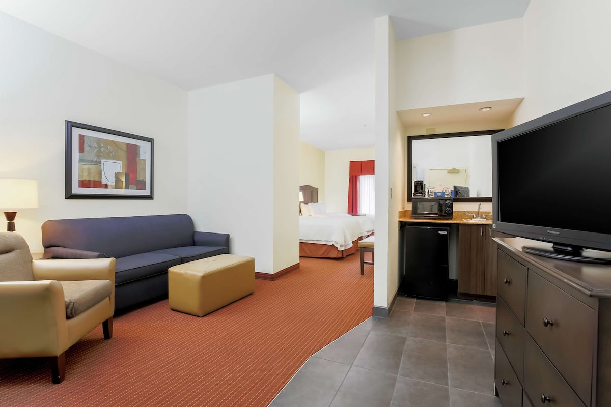 A hotel room with a sofa, armchair, ottoman, a television, a kitchen area with a refrigerator, and a bed in the background.