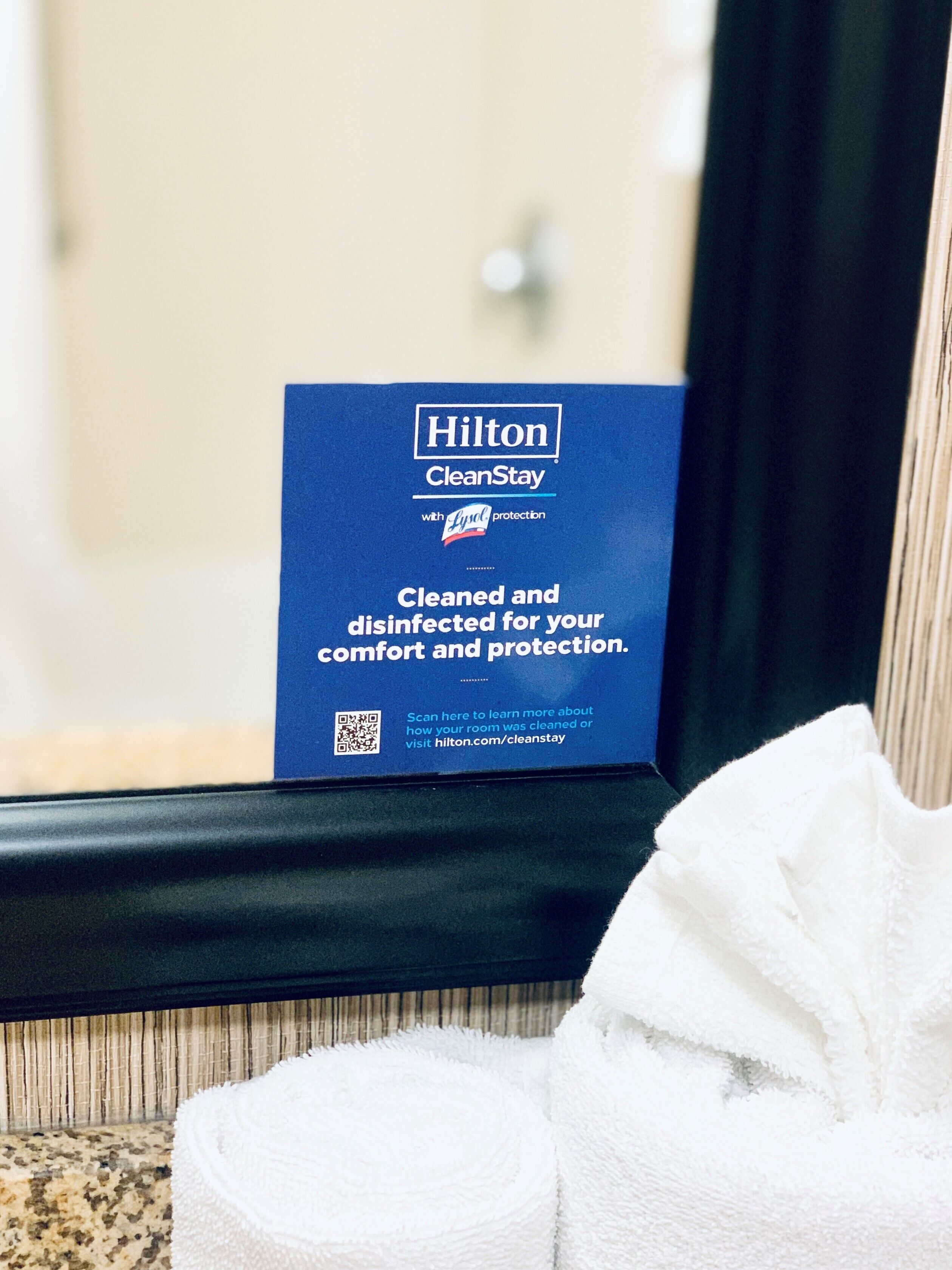 Un letrero azul de Hilton CleanStay con un código QR, colocado sobre un alféizar junto a una toalla blanca.