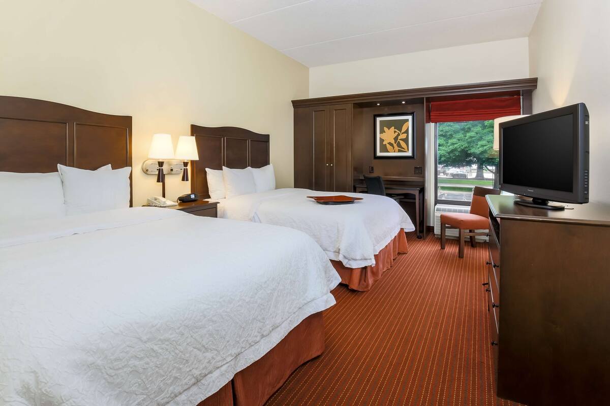 A hotel room with two beds, a desk with a television, and a window with a red valance.