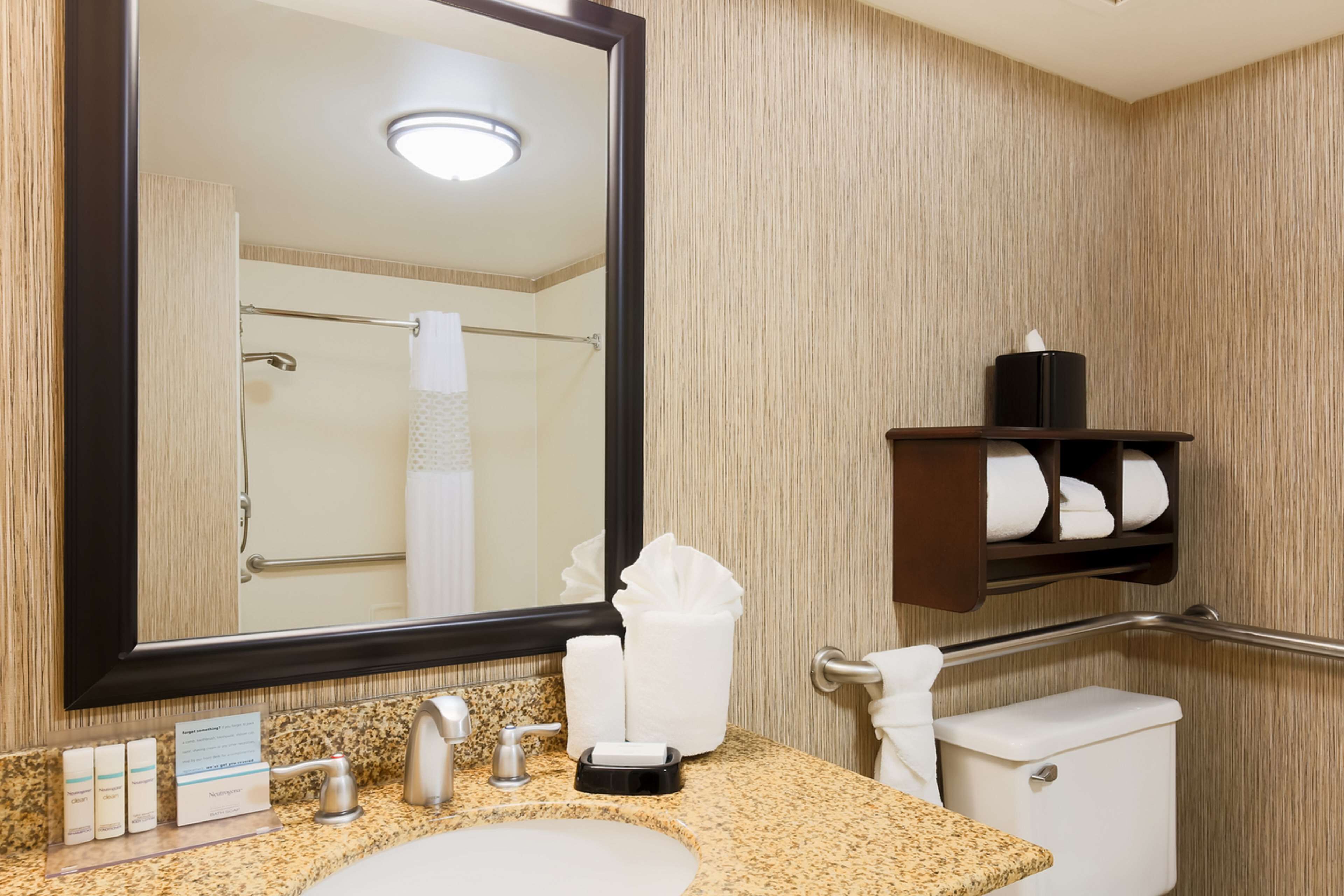 A hotel bathroom with a granite countertop sink, a large mirror, a towel rack, and a shelf with toiletries.