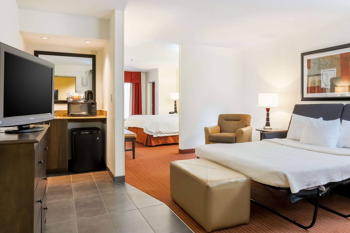 A hotel room with a bed, a TV, a desk with a microwave, a chair, and a small table with a lamp.