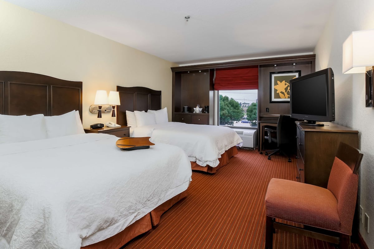A hotel room with two beds, a desk, a chair, a television, and a window with a red curtain.