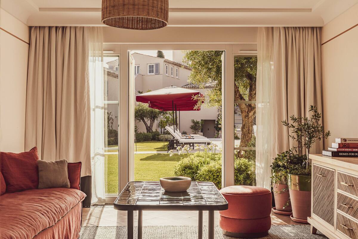 Uma sala de estar moderna com um sofá, uma mesa de vidro com uma tigela, um banquinho vermelho, e vista para um jardim e área de estar externa através de grandes janelas.