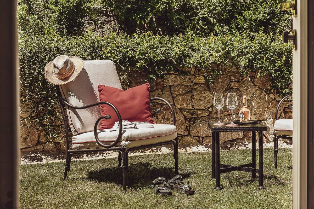Um pátio com jardim, uma cadeira, uma mesa com copos de vinho e uma garrafa, e um muro de pedra.