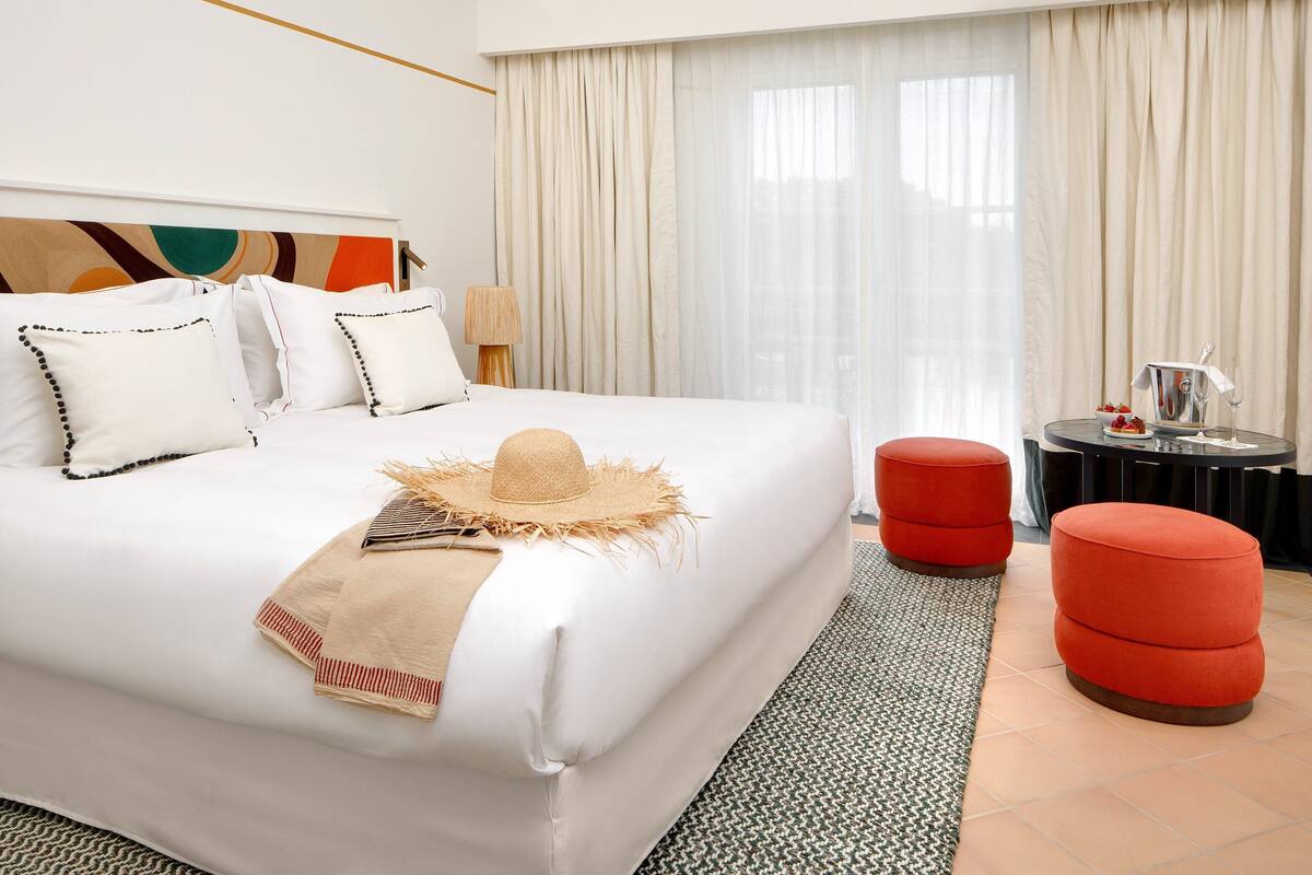 Quarto de hotel com uma cama grande, um banquinho vermelho, uma mesinha com uma bebida e uma janela ampla com cortinas.