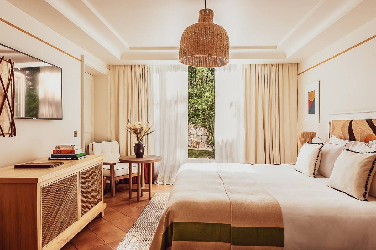 Quarto bem iluminado com uma cama grande, mesa de cabeceira de madeira, televisão e vista para o verde lá fora.
