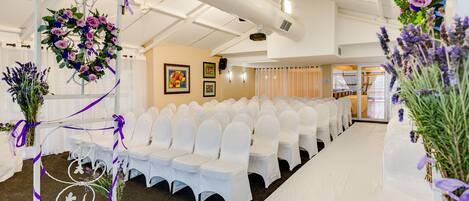 A venue with rows of white chairs, a stage with floral decorations, and a neutral-colored wall with framed pictures.